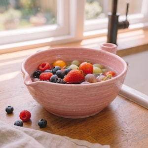Puede incluir: Un cuenco colador de cerámica de color rosa claro con un exterior texturizado y acanalado y un pequeño pico vertedor. El cuenco está lleno de agua y una variedad de productos frescos, incluyendo fresas rojas, frambuesas, moras, uvas y tomates cherry.