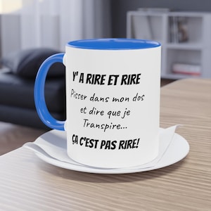 Pode incluir: Uma caneca de cerâmica branca com uma alça e borda interna azul brilhante. A caneca apresenta um texto impresso em preto que diz: Y'A RIRE ET RIRE, Pisser dans mon dos et dire que je Transpire... ÇA C'EST PAS RIRE!