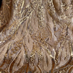 Könnte beinhalten: Ein transparenter Stoff mit einem komplizierten Muster aus goldenen Pailletten und kleinen cremefarbenen Perlen. Das Design besteht aus fließenden, blattartigen Formen und geschwungenen Linien, die eine dichte, strukturierte Oberfläche bilden.