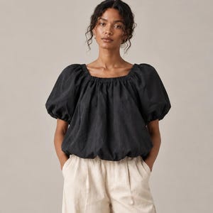 May include: A black blouse featuring a square neckline, gathered elastic detailing, and voluminous puff sleeves. The garment has a relaxed, blouson-style silhouette that gathers at the hem, paired with light beige, high-waisted trousers.