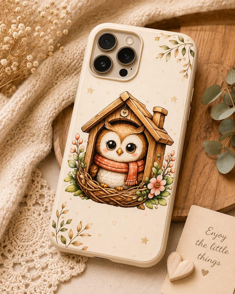 May include: A cream-colored smartphone case featuring a printed illustration of a brown owl wearing a red scarf, sitting inside a wooden birdhouse nestled in a nest with green leaves and pink flowers, accented by small stars and leafy sprigs.