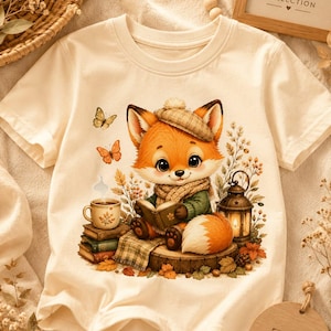 May include: A cream-colored short-sleeve t-shirt featuring a graphic of a fox wearing a plaid beret and scarf, reading a book while sitting on a tree stump. The scene includes a stack of books, a coffee mug, a lantern, and butterflies.