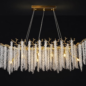 May include: A rectangular gold-toned chandelier featuring a branch-like frame structure. Numerous strands of clear, faceted crystal beads hang vertically from the gold branches, creating a cascading effect. The fixture is suspended by thin wires.