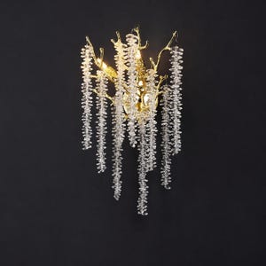 May include: A gold-toned wall sconce with a branch-like design, adorned with cascading clear crystal beads. The fixture has a central light source and is mounted on a dark background, creating a dramatic contrast.