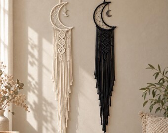 May include: Two crescent moon wall hangings made of knotted cord, one in cream and one in black. Each features a webbed centre, a small hanging moon charm, and long, layered fringe tassels that extend downwards from the crescent frame.
