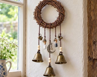 May include: A wall hanging featuring a circular grapevine wreath with five brown cords suspended from it. The cords hold three brass bells, two small glass vials with cork stoppers, wooden beads, and a central metal crescent moon charm.