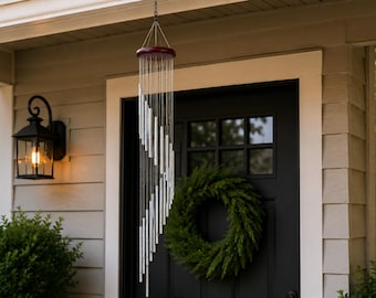 May include: A hanging wind chime with silver tubes and a dark red top. The wind chime is suspended in front of a black door with a green wreath. A black lantern with a lit bulb is mounted on the wall.