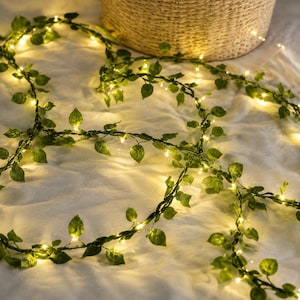 May include: A string of artificial green ivy leaves intertwined with thin copper wire fairy lights that emit a warm yellow glow. The vine features numerous small, pointed leaves distributed along the flexible cord, creating a decorative lighting effect.