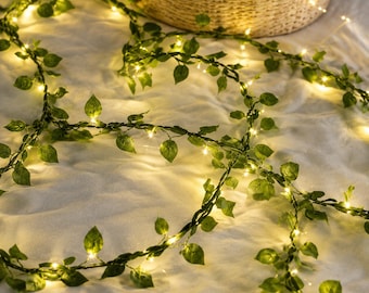 Puede incluir: Una guirnalda de hojas de hiedra artificiales verdes entrelazadas con finos cables de cobre y pequeñas luces que emiten un cálido resplandor amarillo. La enredadera presenta numerosas hojas pequeñas y puntiagudas a lo largo del cordón flexible.