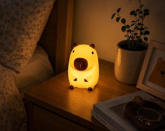 May include: A glowing capybara-shaped night light with a soft yellow illumination, featuring a rounded body, small brown ears, a brown snout, and tiny brown feet, emitting a warm light from its smooth, matte surface.