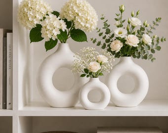 May include: Three matte white ceramic donut-shaped vases of varying sizes arranged on a shelf. The two larger vases hold white hydrangea and white roses with eucalyptus, while the smallest centre vase holds white roses and baby's breath.