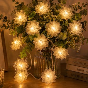 Puede incluir: Una cadena de luces brillantes con forma de flor blanca ilumina un arreglo floral en un jarrón de cristal. Las luces están envueltas alrededor de las flores y la vegetación, creando un ambiente cálido. Una vela y libros están en el fondo.