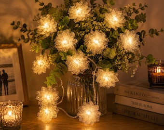 May include: A string of illuminated, white flower-shaped fairy lights decorates a floral arrangement in a glass vase. The lights are wrapped around the flowers and greenery, creating a warm atmosphere. A candle and books are in the background.
