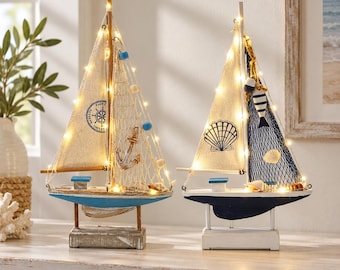 May include: Two decorative model sailboats featuring off-white fabric sails, one with a ship wheel graphic and the other with a seashell graphic. Both boats are adorned with fishing nets, small shells, and warm white string lights.