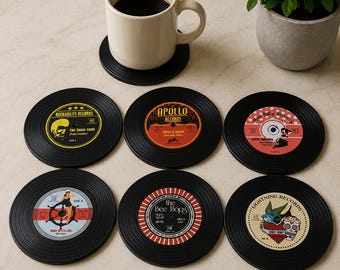 May include: A set of six circular coasters designed to look like black vinyl records with colourful centre labels featuring various graphics and text including Rockability Records, Apollo Records, and The Bee Bops, arranged with a white coffee mug.