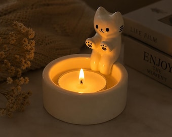 May include: A white ceramic candle holder featuring a small, seated cat figure attached to the rim. The cat has black painted eyes, a nose, and paw pads. A lit tea light candle sits inside the circular base, emitting a warm, golden glow.