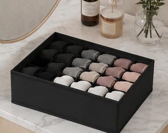 May include: A rectangular black fabric drawer organiser featuring multiple internal compartments. The organiser is filled with rolled socks in a gradient colour palette, transitioning from solid black and dark grey to light grey, beige, and soft pink tones.