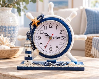 May include: A nautical-themed table clock shaped like a ship's wheel with a white and blue finish. The clock face features black Arabic numerals, ornate black hands, and a red second hand. It is decorated with a lighthouse, a starfish, and rope.