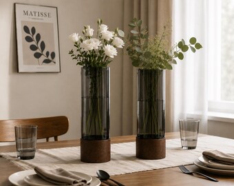 May include: Two cylindrical glass vases with a dark grey tint, each on a wooden base, display white flowers and greenery. The vases are on a table set with plates, glasses, and cutlery. A framed print hangs on the wall.