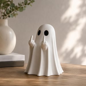 Könnte beinhalten: Eine weiße Keramik-Geisterfigur mit schwarzen Augenlöchern, die mit beiden Händen den Mittelfinger zeigt. Der Geist steht auf einer Holzoberfläche. Eine Vase und Bücher befinden sich im Hintergrund.