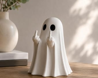 May include: A white ceramic ghost ornament with black eye holes, giving the middle finger with both hands. The ghost is on a wooden surface. A vase and books are in the background.