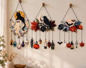 May include: Three decorative wall hangings featuring Halloween themes. The first shows a ghost with a black cat, the second a raven with autumn leaves and a moon, and the third a bat with a spiderweb, pumpkin, and lantern, all suspended by black cords.