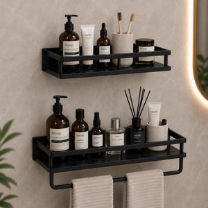 Puede incluir: Dos estantes rectangulares de metal negro montados en la pared con barandillas protectoras. El estante superior contiene botellas ámbar, un tubo y un portacepillos. El estante inferior sostiene botellas, un difusor, un tubo y una barra con dos toallas beige.