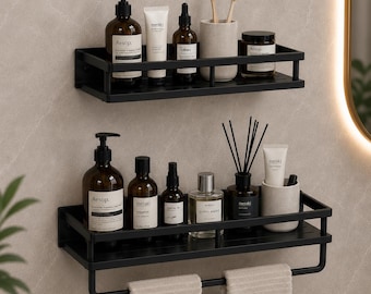 May include: Two rectangular black metal wall-mounted shelves with protective railings. The top shelf holds amber bottles, a tube, and a toothbrush holder. The bottom shelf holds bottles, a diffuser, a tube, and a towel bar with two beige towels.