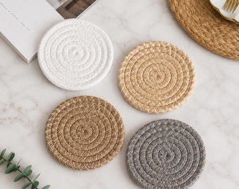 May include: A set of four circular coasters made from coiled rope, featuring a textured spiral pattern. The coasters are presented in four distinct colours: solid white, light tan, medium brown, and dark grey, each with a consistent woven design.