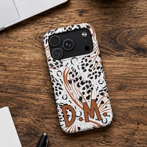 Könnte beinhalten: Eine Smartphone-Hülle mit weißem Hintergrund, einem schwarzen, dalmatinerartigen Punktemuster und braunen, abstrakten Linienakzenten. Die Buchstaben D und M sind in einer fetten braunen Schrift in der Mitte der Hülle aufgedruckt.