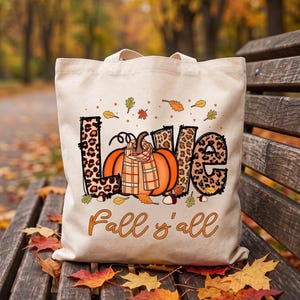 Könnte beinhalten: Eine cremefarbene Stofftasche mit dem Wort LOVE im Leopardenmuster, wobei ein orangefarbener Kürbis mit kariertem Schal den Buchstaben O ersetzt. Darunter steht der orangefarbene Schriftzug Fall y'all, umgeben von kleinen Herbstblättern.