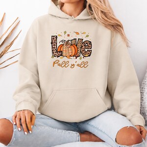 Könnte beinhalten: Ein beigefarbener Kapuzenpullover mit einem Grafikdruck des Wortes LOVE im Leopardenmuster, das einen orangefarbenen Kürbis mit kariertem Schaldesign enthält, umgeben von kleinen Herbstblättern und dem Text Fall y'all darunter.