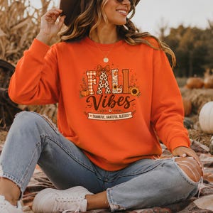 Könnte beinhalten: Ein orangefarbenes Sweatshirt mit Rundhalsausschnitt und einem Grafikdruck mit dem Text FALL VIBES sowie THANKFUL, GRATEFUL, BLESSED. Das Design zeigt einen Kürbis, eine Sonnenblume sowie Karomuster und Leopardenmuster auf dem orangefarbenen Stoff.