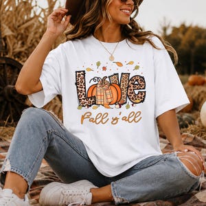 Könnte beinhalten: Ein weißes Kurzarm-T-Shirt mit einem Grafikdruck, der das Wort LOVE in Leopardenmuster, einen orangefarbenen Kürbis mit kariertem Schal und den Text Fall y'all in orangefarbener Schreibschrift zeigt, verziert mit kleinen Herbstblättern.