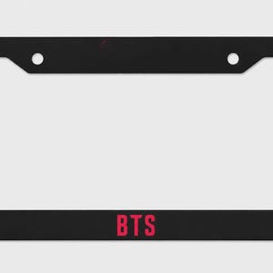 May include: Black license plate frame with the letters "BTS" in red at the bottom. The frame has two holes at the top for mounting. The background is plain white.