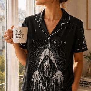 May include: A black short-sleeve button-down pajama shirt and matching shorts set featuring white piping and a graphic print of a hooded figure with the text Sleep Token. The fabric includes celestial patterns, stars, and a crescent moon design.