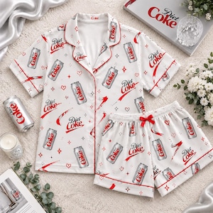 May include: A white short-sleeve button-down pajama shirt and matching shorts set featuring an all-over print of Diet Coke cans, red hearts, and four-pointed stars. The set includes red piping trim and a red ribbon bow on the shorts waistband.