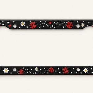 May include: A rectangular black license plate frame featuring a repeating pattern of red ladybugs, white daisies, small white polka dots, and tiny red hearts. The design includes thin dashed lines that trail behind the ladybugs across the frame.