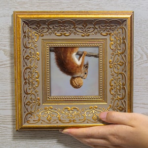 Puede incluir: Una pintura cuadrada de una ardilla roja colgada boca abajo de una rama mientras sostiene una nuez marrón, encerrada en un marco dorado ornamentado y texturizado con un borde interior beige y patrones florales en relieve en los bordes.