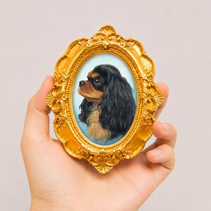 Puede incluir: Un marco ovalado de color dorado con detalles florales en relieve que rodea el retrato de un perro Cavalier King Charles negro y fuego sobre un fondo azul claro, presentado como un pequeño objeto decorativo sostenido en la mano.