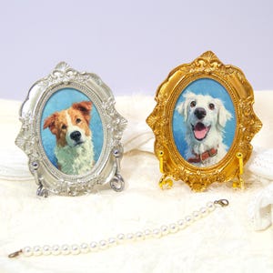Puede incluir: Dos marcos de fotos ovalados, uno plateado y otro dorado, cada uno con un borde ornamentado en relieve. El marco plateado contiene el retrato de un perro marrón y blanco, mientras que el dorado muestra un perro blanco con collar rojo. Una pulsera.