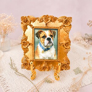 Puede incluir: Un marco de fotos rectangular de color dorado con detalles florales texturizados y ornamentados, apoyado sobre un soporte de metal dorado a juego. El marco contiene el retrato de un bulldog marrón y blanco con un collar azul y una placa.