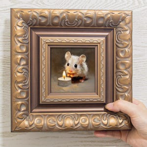 Puede incluir: Una pequeña pintura cuadrada en un marco ornamentado de color bronce metálico. La obra de arte representa un ratón blanco con orejas grandes sentado junto a una vela de té encendida con una llama visible, sobre un fondo de tonos neutros.