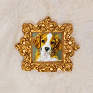 Puede incluir: Una pintura al óleo cuadrada de un cachorro de spaniel marrón y blanco con grandes ojos oscuros, colocada dentro de un marco dorado ornamentado con intrincados adornos y tallas en forma de hoja en los bordes, creando un aspecto decorativo.