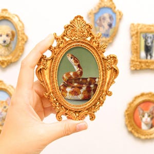 Può includere: Una cornice portafoto ovale di colore dorato con un design ornamentato che racchiude un dipinto dettagliato di un serpente con motivi marroni e bianchi. La cornice presenta intricati decori e intagli floreali lungo i bordi esterni.