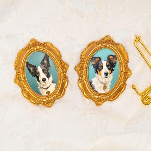 Puede incluir: Dos marcos ovalados dorados y ornamentados con retratos de perros sobre un fondo verde azulado. El marco izquierdo contiene un perro blanco y negro con una placa de corazón, y el derecho un perro tricolor con una placa. Un soporte de metal dorado.