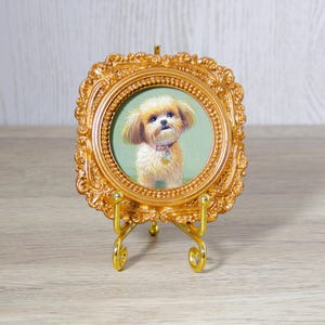 Puede incluir: Un pequeño marco cuadrado de color dorado ornamentado con un borde interior circular. Dentro del marco hay una pintura de un perro esponjoso de color marrón claro con un collar rosa. El marco se sostiene sobre un soporte de metal dorado.
