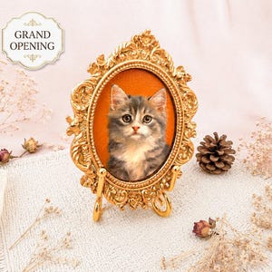 Puede incluir: Un marco ovalado de color dorado ornamentado que contiene el retrato de un gatito gris y blanco sobre un fondo naranja sólido. El marco presenta volutas intrincadas y se apoya en un pequeño soporte dorado. El texto GRAND OPENING es visible arriba.