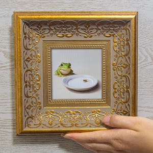 Puede incluir: Una pieza de arte mural cuadrada que presenta una pintura de una rana verde sentada junto a un plato blanco con una pequeña abeja. La obra está enmarcada en un marco dorado ornamentado y texturizado con patrones en relieve y borde beige.