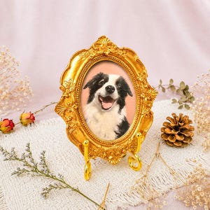 Puede incluir: Un marco de fotos ovalado de color dorado con detalles florales y volutas en relieve. El marco se apoya en un soporte de metal dorado a juego y contiene el retrato de un perro blanco y negro con una expresión alegre.